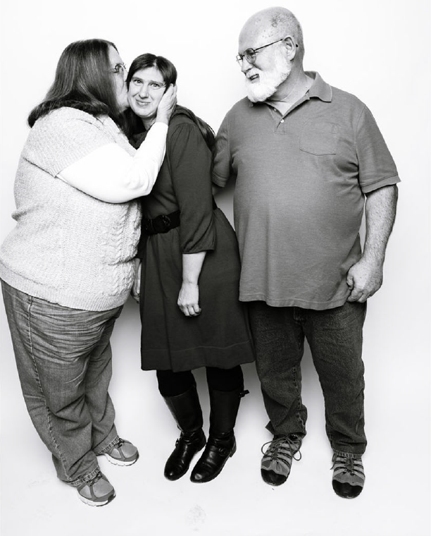 A black-and-white photo of three people standing together against a plain background. A woman on the left is kissing the cheek of a younger woman in the middle, who is smiling. A man on the right, wearing glasses, is also smiling and looking at the women.