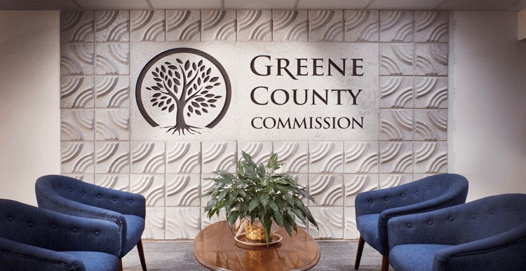 A modern office waiting area features four blue upholstered chairs around a round wooden table with a potted plant. The back wall displays the text "Greene County Commission" next to a logo of an intricate tree with a circular border, set against a tiled backdrop.